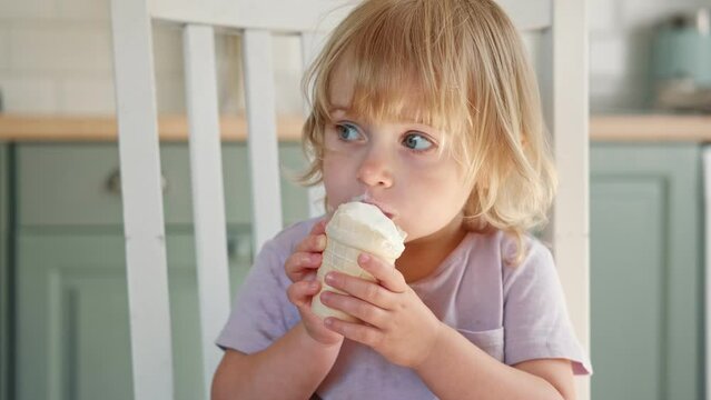 Baby girl enjoying ice cream. Pretty little toddler eating an ice-cream indoors, at home. Dining room background. Small child eats plombir and cream messy on her mouth. Cute kid with tasty sweet food