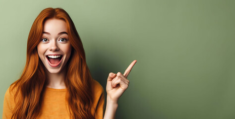 An excited young ginger teenage girl smiling and pointing her index finger up at the copy space expressing wow emotion standing isolated on green background studio copy space, advertisement concept