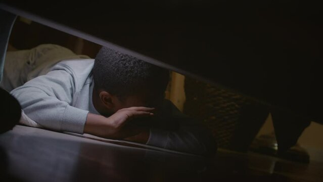 Slowmo of frightened African American boy in pajamas using flashlight while looking for monsters under his bed late at night after watching horror film