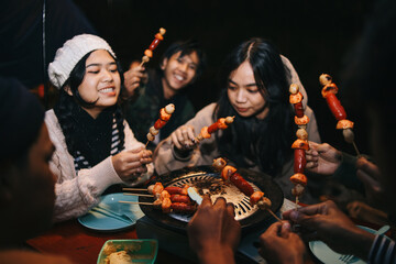 Happy young Asian friends holding sausage and meat skewers in barbeque party