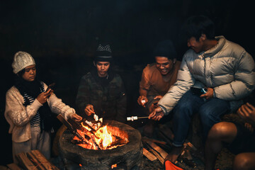 Group of Asian friends gather around campfire and grilling marshmallows on sticks during vacation in the camp site