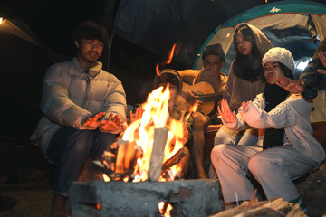 Group of Asian friends sitting around campfire, playing guitar and singing songs while warming hand...
