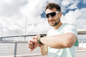 Uses the fitness watch app. A sporty man in a T-shirt and glasses for running training in the open air. Happy guy hobby running instructor before exercises.