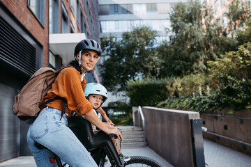 Mother carring her son on a child bike carrier, seat, both wearing helmets. Mom commuting with a...