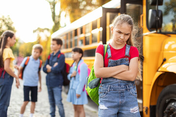 Upset girl standing alone near school bus while classmates chatting on background