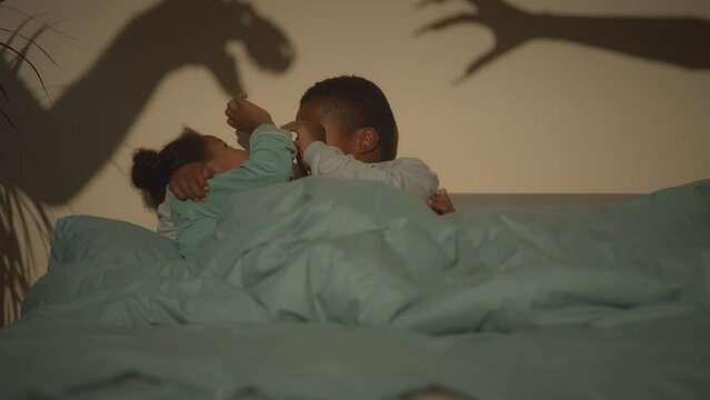Slowmo of two African American tween siblings lying together under blanket in bed at dark room and screaming with scary shadows on wall playing their game