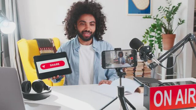 Young popular Arabian man leading online blog or tv show calls for sharing video and demonstrates tablet computer with request for subscribers sits at table with phone and microphone in home office.