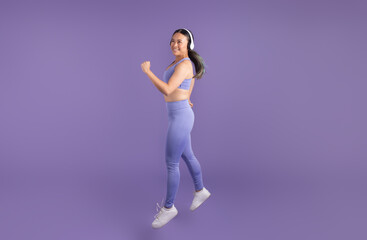 Fototapeta na wymiar Asian woman in headphones jumping over purple studio backdrop, full length