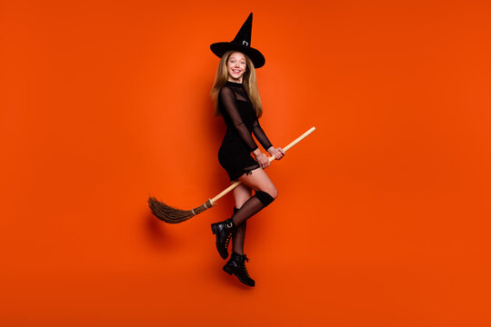Full length photo of lovely blonde teen lady sit broom flying dressed black halloween garment isolated on orange color background