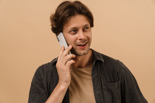 Smiling man talking on mobile phone while standing isolated over beige wall