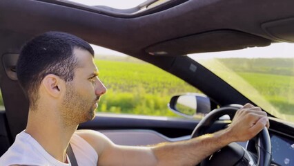 Profile of young handsome man driving a modern electric car with sunlight at background. Young male driver riding on electrical vehicle at countryside. Concept of journey and trip. Close up