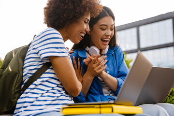 Two joyful female students having fun while using laptop outdoors in campus