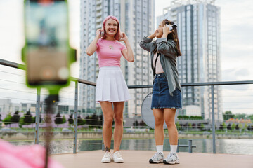 Two smiling girls making video with smartphone while standing on city waterfront