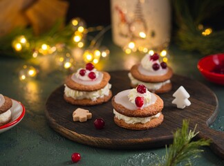 Christmas sandwich cookies filled with whipped cream and sugar icing, decorated with cream and berries, on a brown wooden board on a green concrete background.