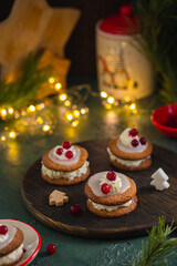 Christmas sandwich cookies filled with whipped cream and sugar icing, decorated with cream and berries, on a brown wooden board on a green concrete background.