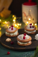 Christmas sandwich cookies filled with whipped cream and sugar icing, decorated with cream and berries, on a brown wooden board on a green concrete background.