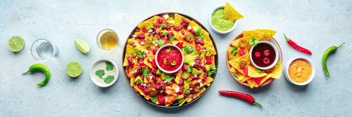 Loaded nachos panorama. Mexican nacho chips with beef, overhead flat lay shot with guacamole sauce, cheese salsa, tequila drinks, limes, and chili peppers - obrazy, fototapety, plakaty