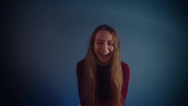 Portrait of a young beautiful laughing happy smiling pretty caucasian girl with long brown hair in a burgundy sweater looking at the camera on a blue background. Emotion of joy and happiness