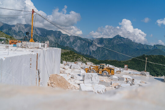 Blocks of cut white Carrara marble in the quarry in front of the rock face ready for transportation in Tuscany Italy. Quarry view with heavy front loader moving rubble debris. Landscape mountains.