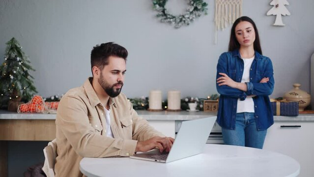Distressed couple experiencing tension in the decorated kitchen. Upset woman avoiding conversation with her husband in a private home setting. Guilty man focuses on laptop during Christmas time.