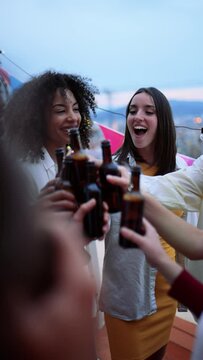Diverse group of carefree friends having fun and drinking beer in a rooftop party in a city. Multicultural people celebrating and toasting with bottles at evening in a terrace