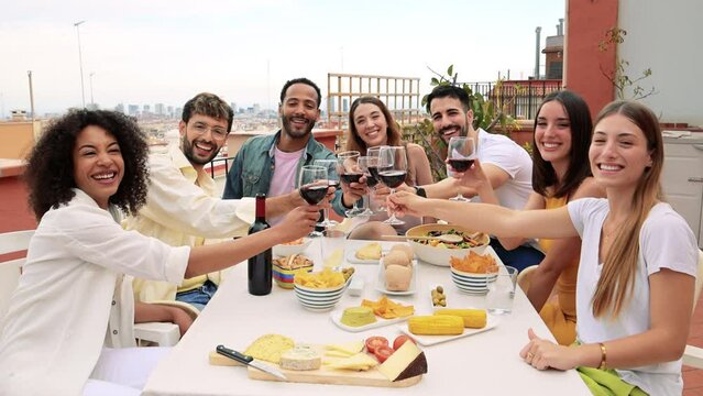 Happy Group Of Friends Toasting With Wine Glasses In A Terrace Looking At Camera. Diverse Group Of Young People Drinking In A Rooftop Dinner Staring At Camera.