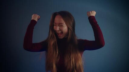 Portrait of a young beautiful smiling happy jubilant pretty caucasian girl with raised hands, long brown hair in a burgundy sweater on a blue background. Emotions of triumph, joy and jubilation