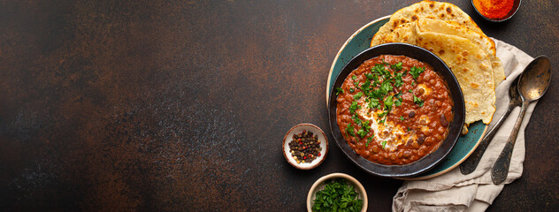 Traditional Indian Punjabi dish Dal makhani with lentils and beans in black bowl served with naan flat bread, fresh cilantro and two spoons on brown concrete rustic table top view. Space for text. - obrazy, fototapety, plakaty