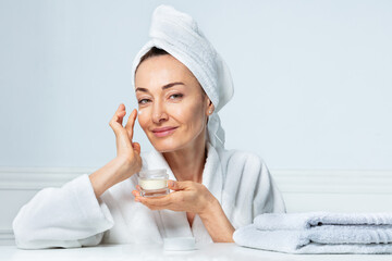 Woman in turban smiling, applying face cream at bathroom - obrazy, fototapety, plakaty