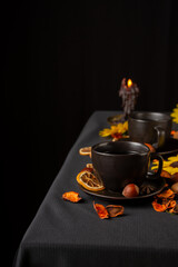 Aerial view of two cups of tea on table with black tablecloth with dried fruits, yellow flowers and dark lit candle, black background, vertical, with copy space