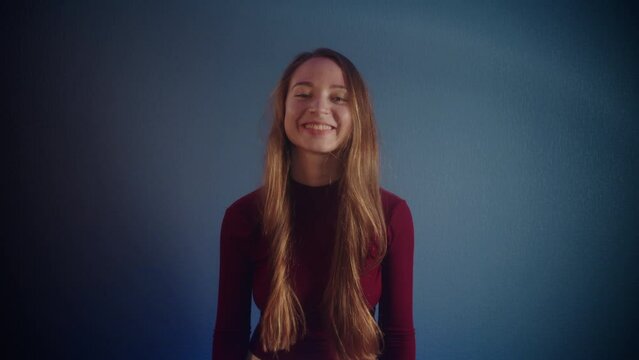 Portrait of a young beautiful playful agreeing smiling pretty caucasian girl with long brown hair in a burgundy sweater looking at the camera and shaking her head on a blue background