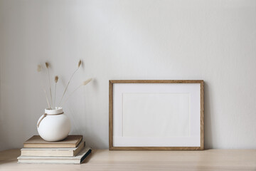 Horizontal blank wooden picture frame mockup on table. Trendy round ceramic vase with dry bunny tails, Lagurus ovatus grass and old books. White wall background. Scandinavian interior, home office.