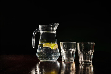 refreshening full jug of fresh and clear water with lemon and empty glasses on black backdrop