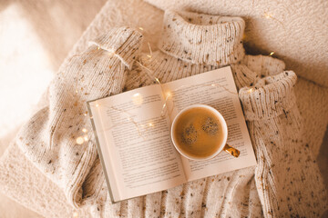 Cup of fresh coffee on open paper book with knitted sweater clothes in chair closeup. Hygge cozy home atmosphere. Top view.