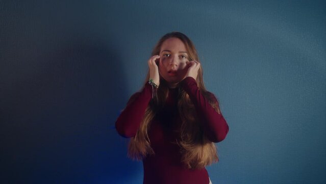 Portrait of a young beautiful erotic attractive caucasian girl in a burgundy sweater looking at the camera on a blue background. She seductively touching her long brown hair and head in a slow motion