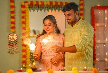 Joyful young indian couples playing with sparklers or firecrackers during diwali festival celebration at home - concept of special occasion, family bonding and traditional culture.