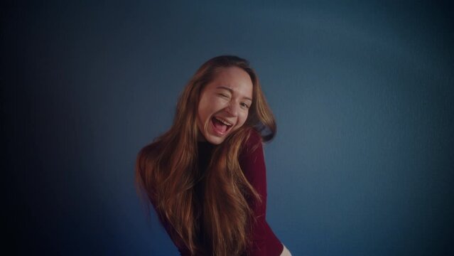 Portrait of a young beautiful playful funny smiling pretty caucasian girl with long brown hair in a burgundy sweater looking at the camera and artistically winking on a blue background