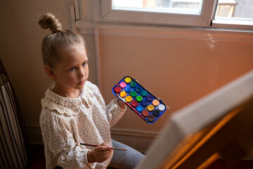 little cute girl draws with paints on canvas at the easel