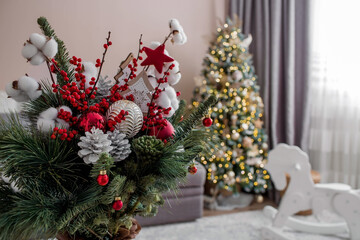 A bouquet of fir trees with red berries, cotton, toys on the table against the background of Christmas tree garlands. Winter decorations in the interior. The concept New Year holidays 2024.