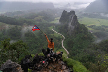 Tourist holding Lao flag on the Nam Xay Viewpoint, During the rainy season in Vang Vieng, Laos. - obrazy, fototapety, plakaty