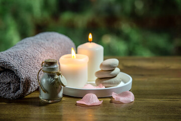 Fototapeta na wymiar spa still life with candles, towel and white stones against green nature background on the wooden table