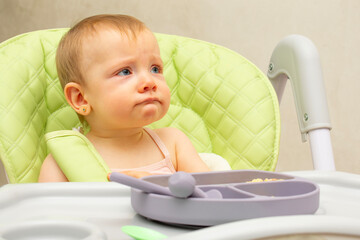 Baby wearing knitted sweater sitting in high chair and feels hungry, holding spoon and eating puree or porridge, enjoying apples and water in yellow bottle, posing isolated on white background.