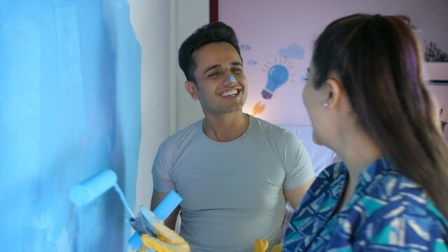 A young man and a pretty woman painting a wall together - rubber gloves for paint work  teamwork  home renovation  couple bonding  couple goals. A cheerful lady with a roller brush teasing her husb...