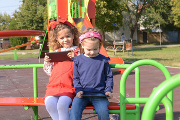 Fototapeta na wymiar Two little girls use a tablet on the playground. Small children playing outdoors