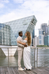 International wedding. The handsome dark-skinned groom in a white suit with lightened hair embraces a beautiful Caucasian bride with a gorgeous hairstyle against the backdrop of Singaporel city 