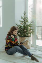 Smiling joyful teenage girl writing wishes in notebook while sitting near Christmas tree at home during winter holidays on New Year's Eve. Soft focus