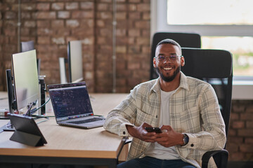 A young African American entrepreneur takes a break in a modern office, using a smartphone to...