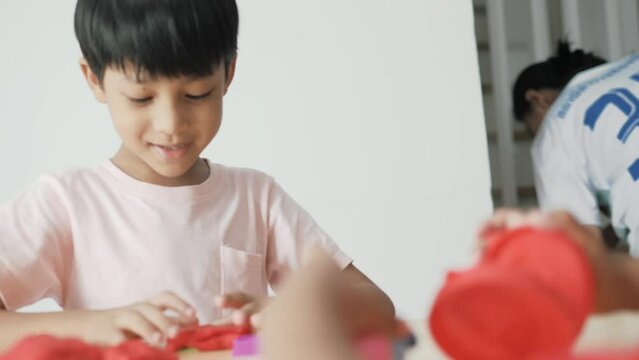 An adorable 4-6 year old Asian boy wearing a pink shirt is happily playing with clay and has many toys placed on a table inside his house. Behind him is a TV and shelves and plants.