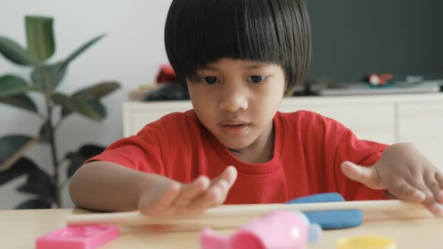 An adorable 3-5 year old Asian boy wearing a red shirt is happily playing with clay and has many toys placed on a table inside his home. Behind there is a TV and a shelf and a plant.