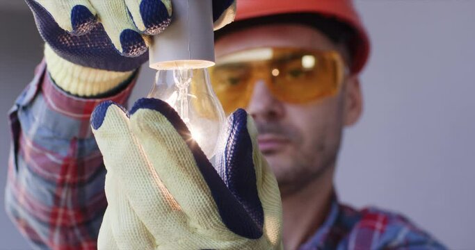 Connect the lighting by screwing in a light bulb. An electrician wearing safety glasses and a hard hat screws a light bulb into a socket.